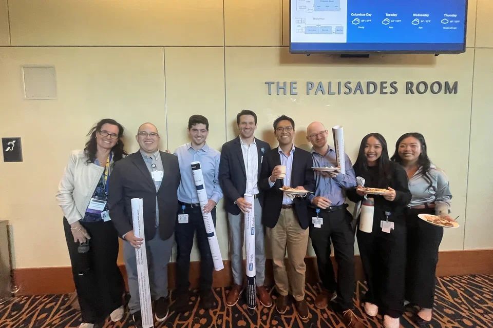 Eight colleagues smiling and holding posters in front of "The Palisades Room" after presenting at DOM Research Day